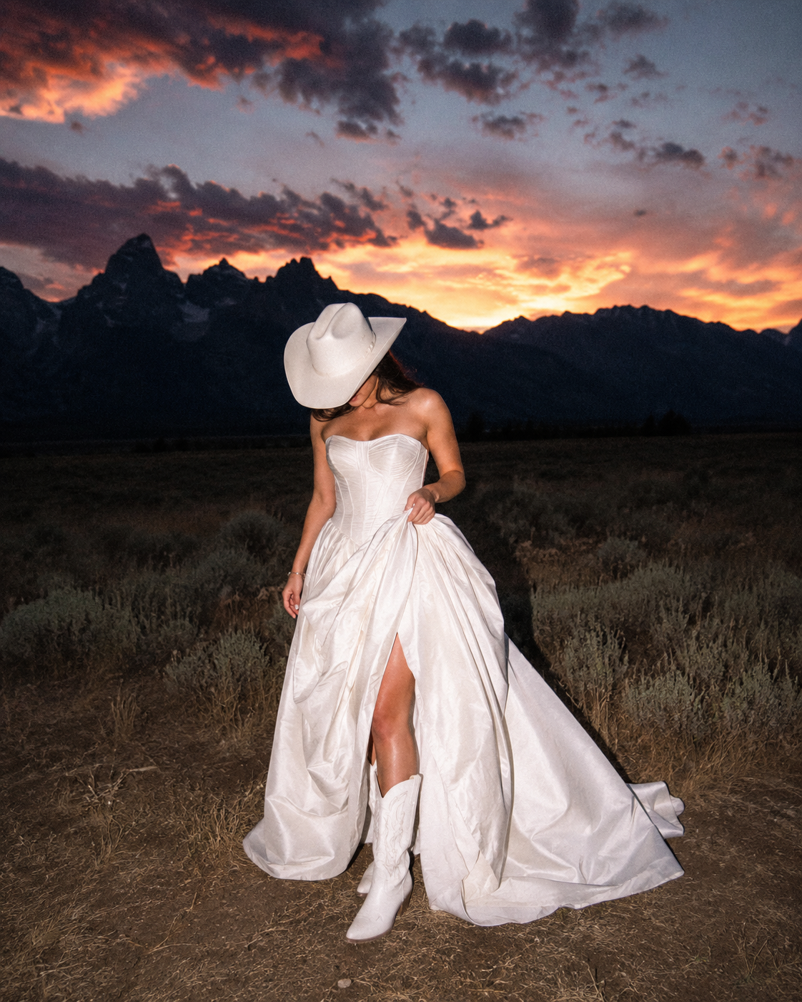 Bride in a white gown and cowboy boots at sunset in front of mountains